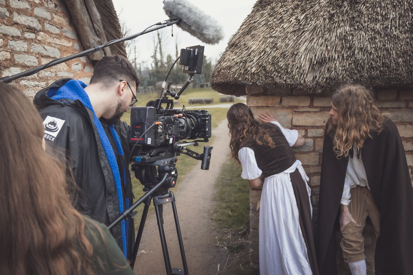 Filmmaker operates camera on tripod filming actors by brick wall.